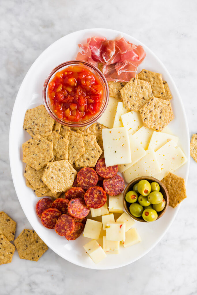 Overhead picture of an oval snack board with spicy deli meats, crackers, cheeses and olives. 