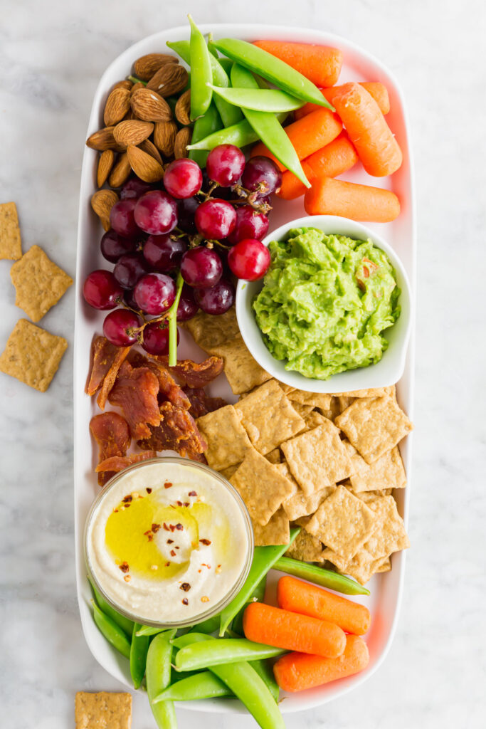 Overhead picture of protein-power snack board rectangle snack board with grapes, carrots, snap peas, almonds, guacamole, hummus and crackers