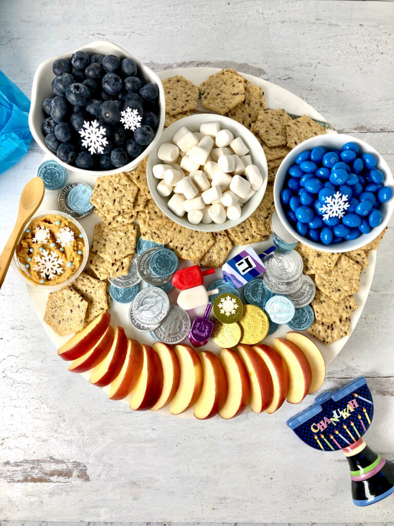 Picture of Hanukkah themed snack board with crackers, marshmallows, blue M&M, apples slices, blueberries and more.